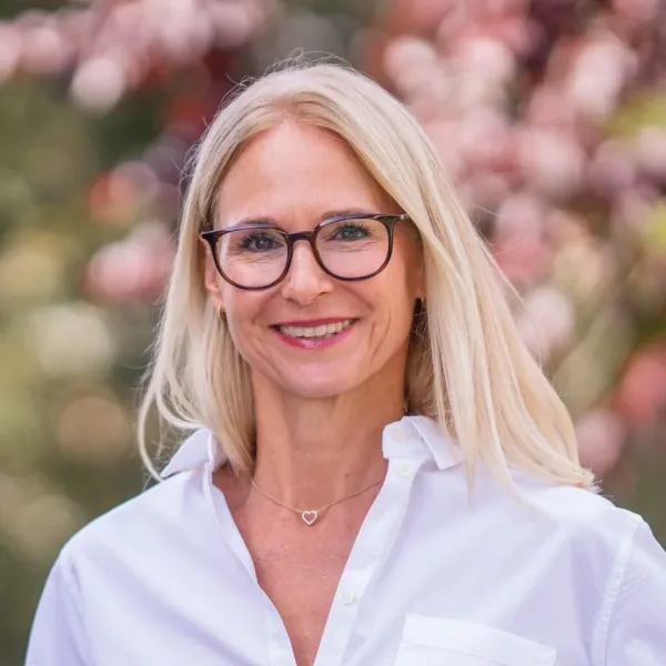 Frau mit Brille lächelt, trägt weißes Hemd, verschwommener Hintergrund.