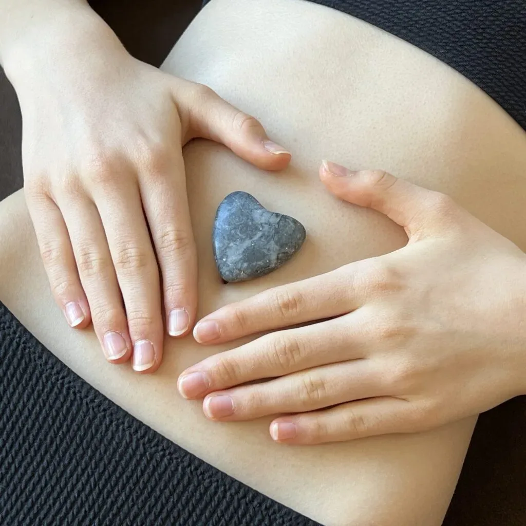 Hände auf Bauch mit herzförmigem Stein, Entspannung und Achtsamkeit symbolisierend.